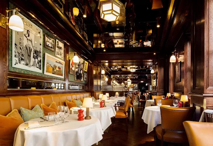 A cozy, elegant restaurant with brown leather chairs, round tables with white tablecloths, warm lighting, and framed photos on dark wood-paneled walls. The atmosphere is inviting and upscale.