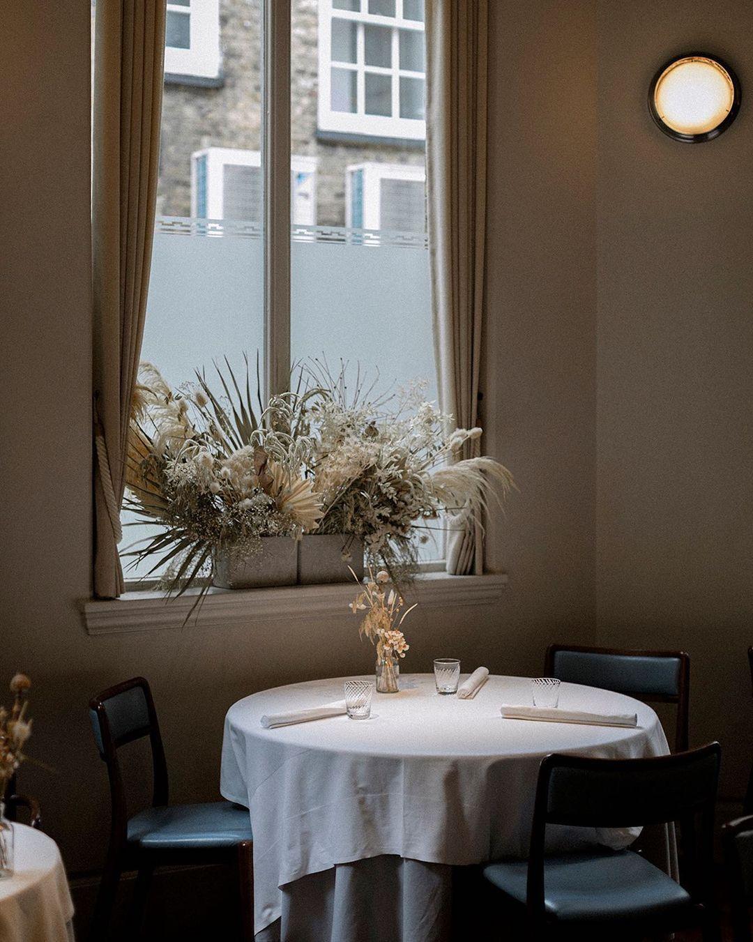 A cosy dining area with a round table covered in a white tablecloth, set with two chairs. The table holds a small vase of dried flowers. A large window with beige curtains and a dried flower arrangement provides natural light. A wall lamp is illuminated at The Clove Club, Shoreditch.