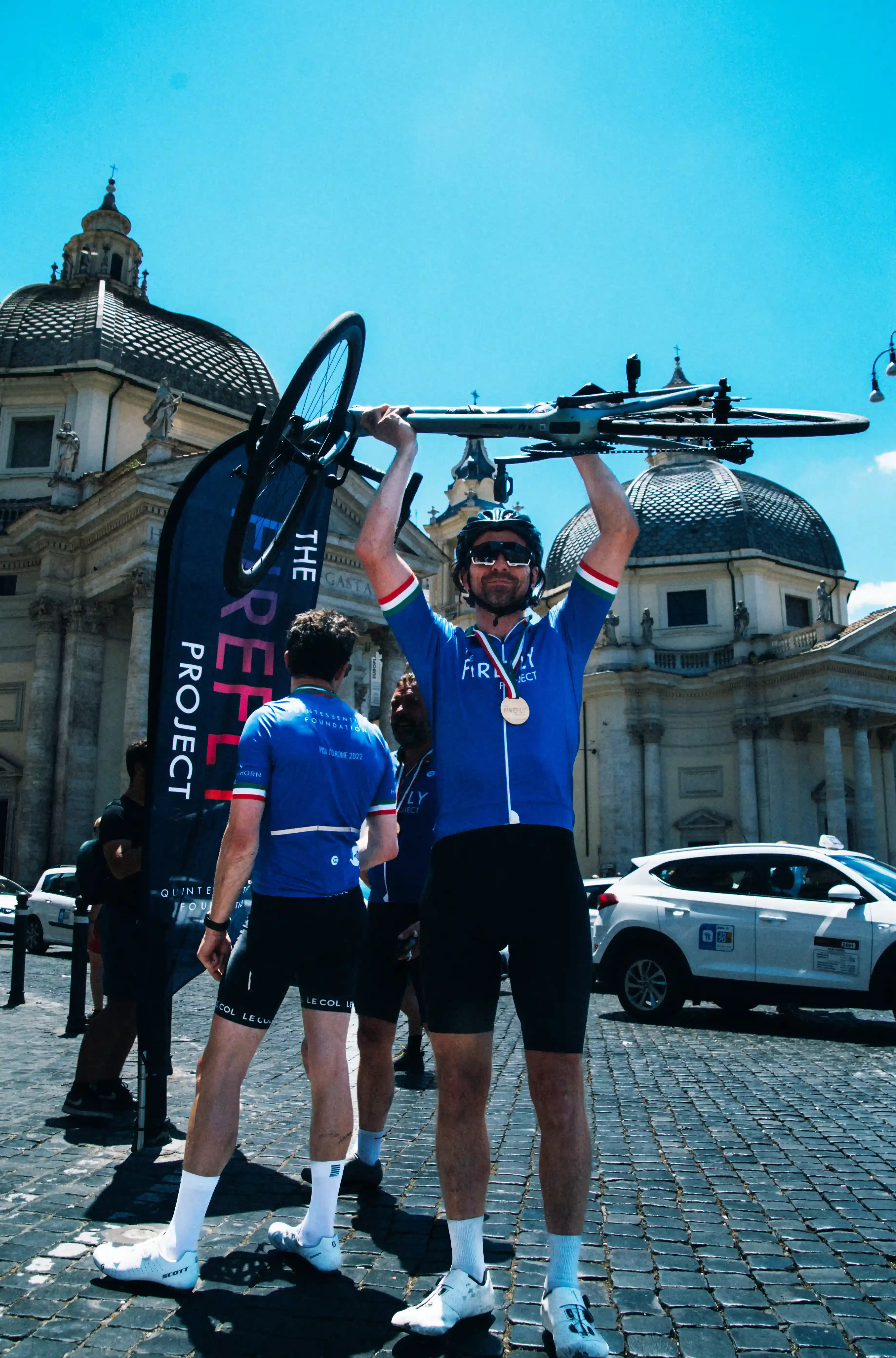 Quintessentially Foundation | The Firefly Project | Man in cycling uniform lifting bike in the air celebrating