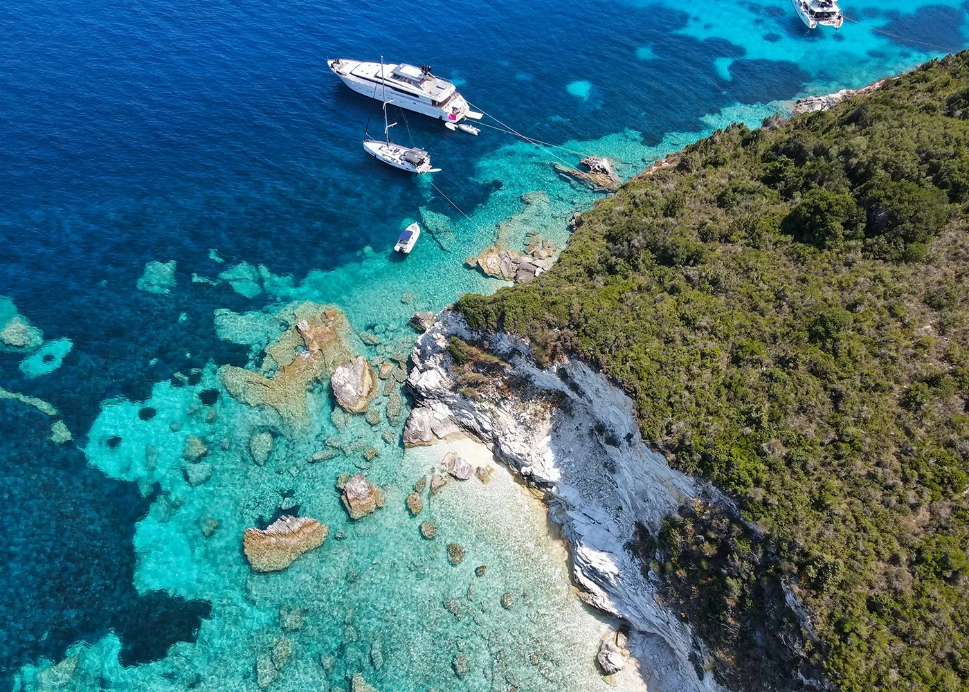Aerial view of a rocky coastline with lush green vegetation on the right. Crystal-clear turquoise waters display visible rocks beneath the surface. Several boats, including a large yacht, are anchored near the shore.