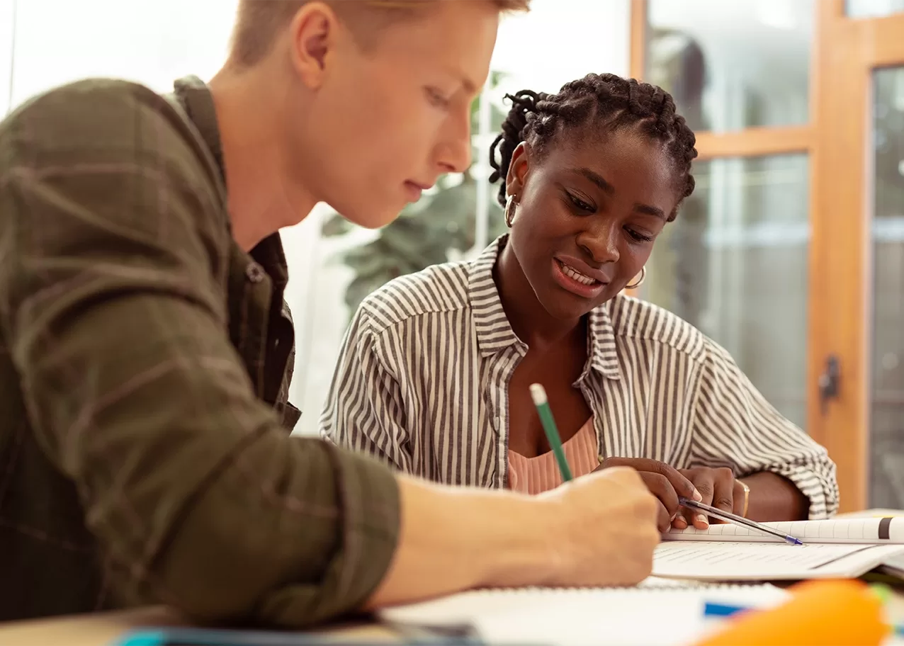 A young man and woman are sitting at a table, working together on notebooks. The man is writing, while the woman smiles and looks on. They appear to be in a study or collaborative session, with books and stationery around them.