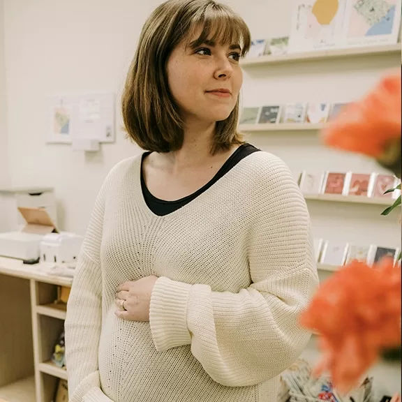 A woman with shoulder-length brown hair and bangs, wearing a cream-colored sweater, stands indoors near a display of cards and shelves, gently resting her hand on her stomach. Orange flowers are visible in the foreground.