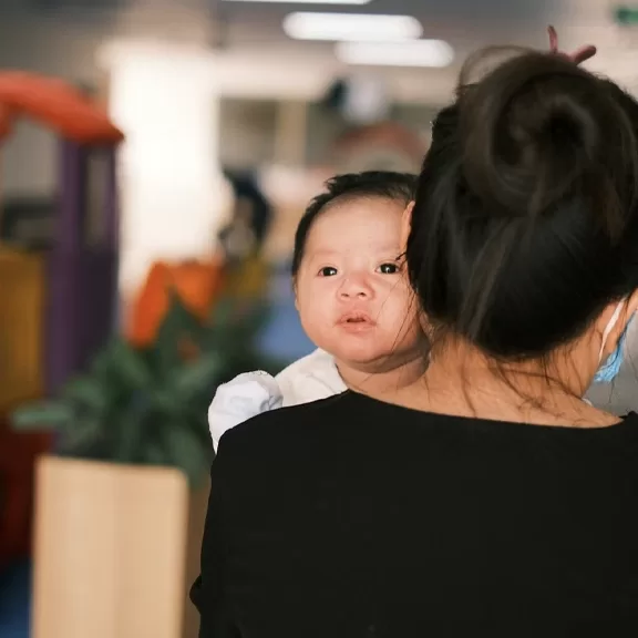 A baby with a curious expression is being carried by an adult with their back to the camera. The background is softly blurred, showing indoor plants and colorful objects.