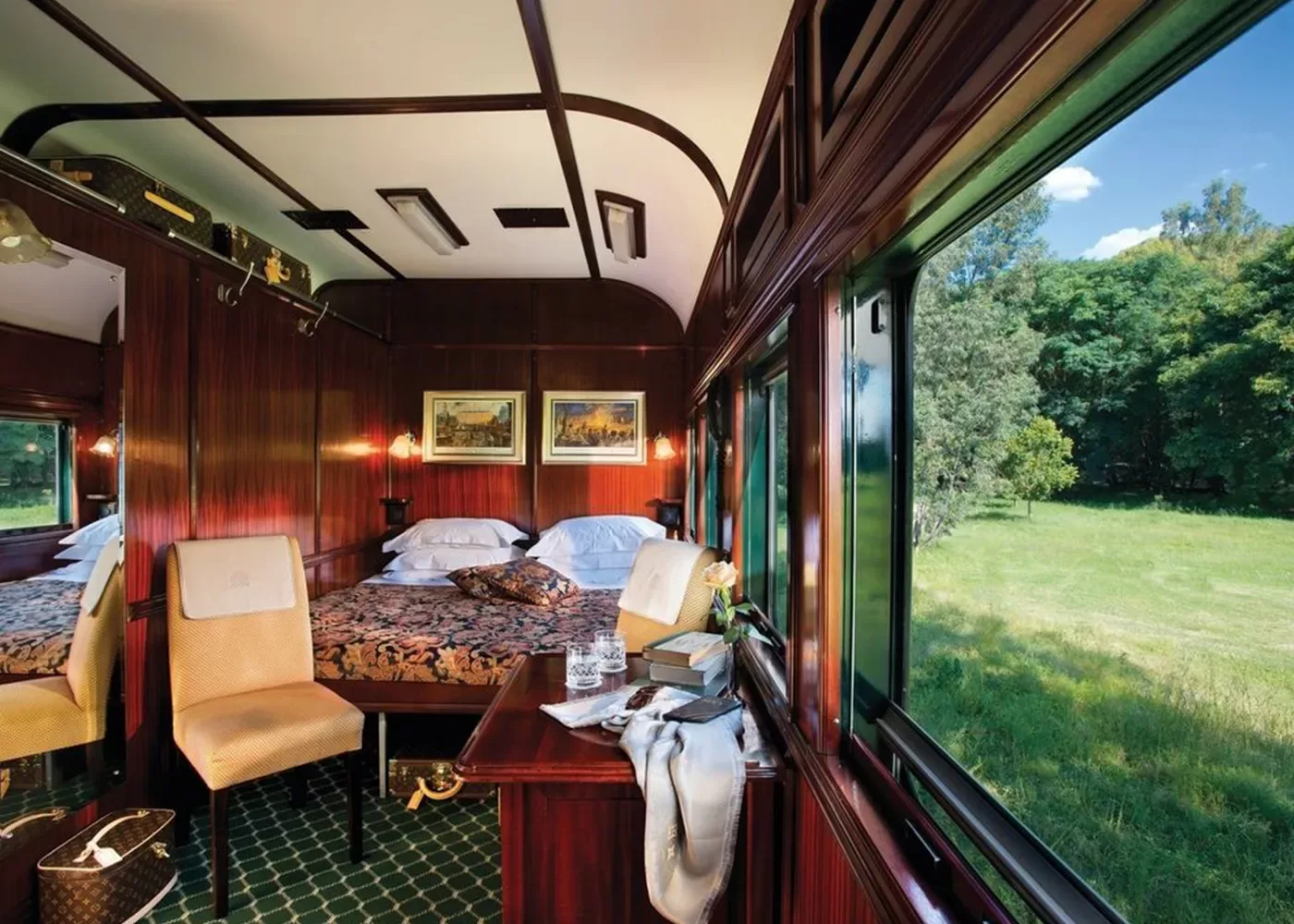 Luxurious train cabin interior aboard The Southern Cross by Rovos Rail in Africa, with wood panelling, featuring a bed with floral bedding, chairs, and a desk with books and a fruit bowl. A large window offers a view of lush greenery and blue sky outside.