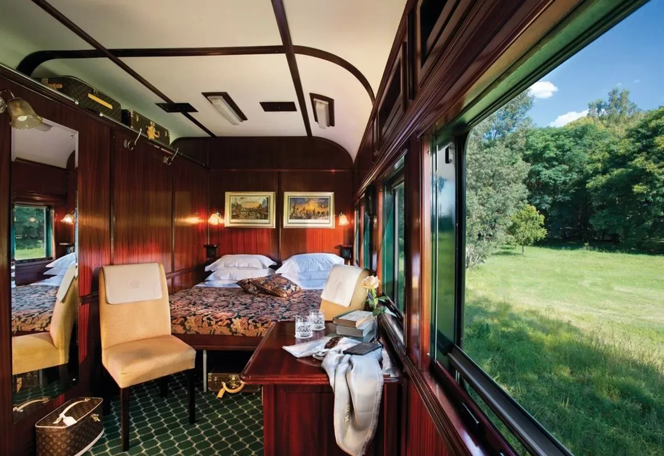 Luxurious train cabin interior aboard The Southern Cross by Rovos Rail in Africa, with wood panelling, featuring a bed with floral bedding, chairs, and a desk with books and a fruit bowl. A large window offers a view of lush greenery and blue sky outside.