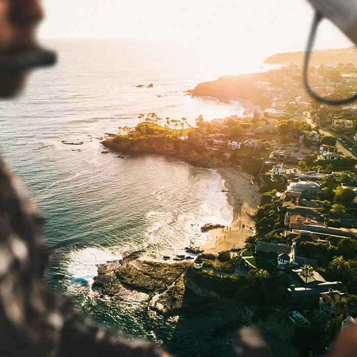 Aerial view of a coastline at sunset, showing waves, sandy beaches, rocky areas, and houses along the shore. Part of a person wearing a headset is visible in the blurry foreground.