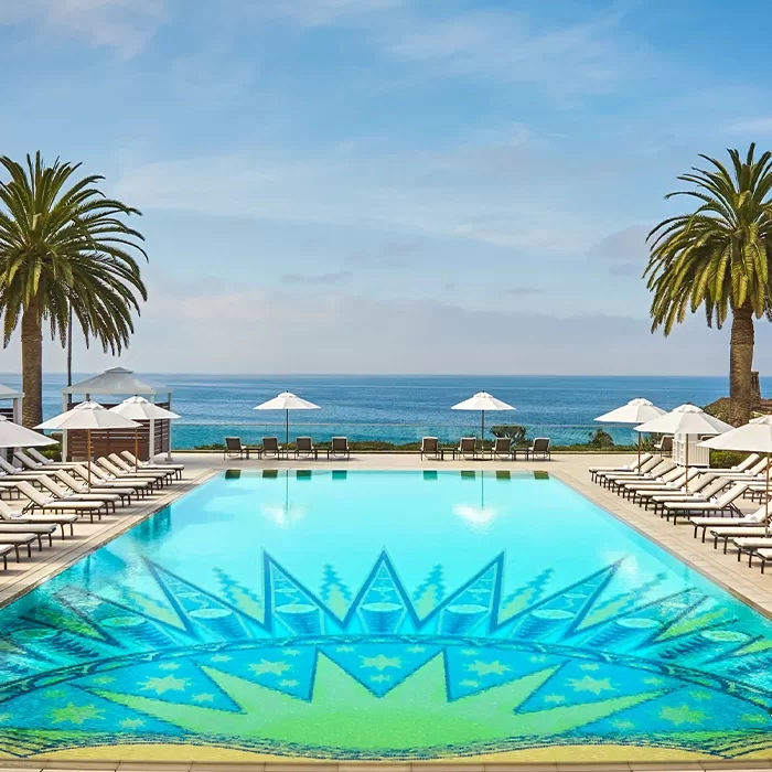 A luxurious pool with a geometric mosaic design is surrounded by lounge chairs and umbrellas, with palm trees on each side and an ocean view in the background under a clear blue sky.