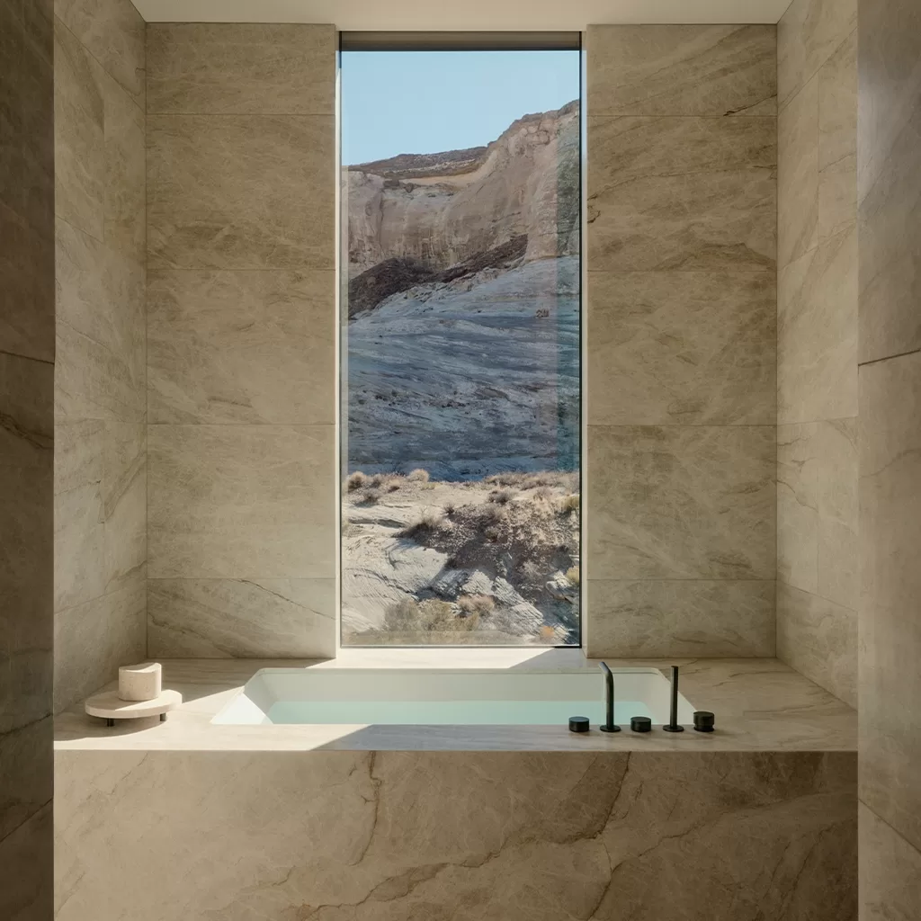 A sunlit bathtub with beige marble surround sits beneath a tall window, offering a view of rocky, arid hills outside. Minimalist bath fixtures and a tray with a cup complete the serene, modern bathroom scene.