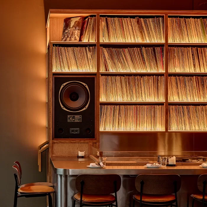 A warmly lit bar with shelves filled with vinyl records, a large speaker, and a countertop set with glasses and napkins. Three chairs are placed in front, creating a cozy, retro atmosphere.