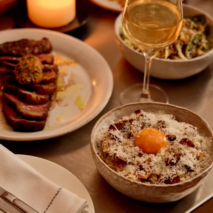 A bowl of pasta topped with grated cheese and an egg yolk sits on a table next to a glass of white wine, a plate of sliced steak with sauce, and a bowl of salad, with a lit candle in the background.