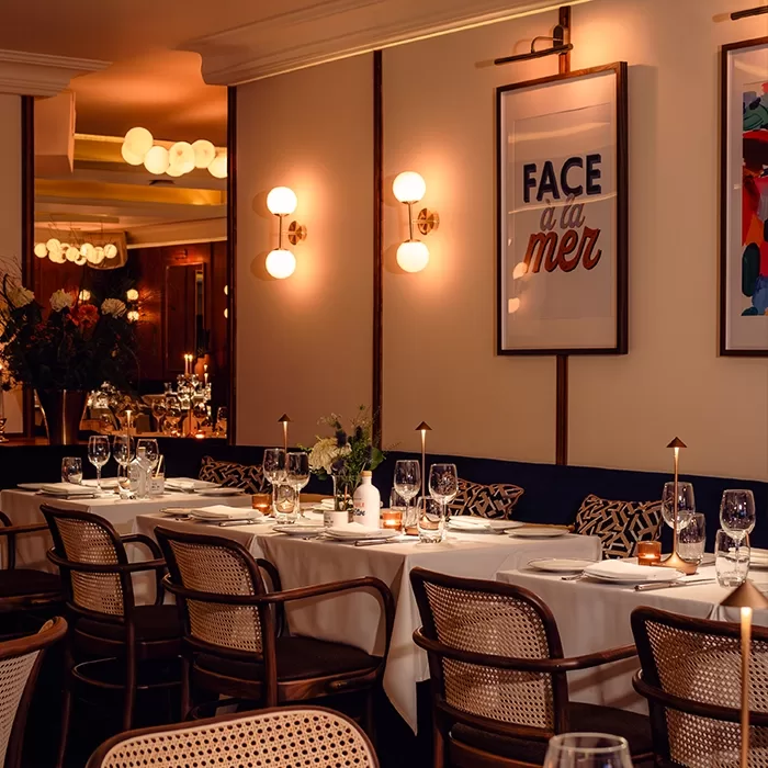 A warmly lit restaurant interior with white tablecloths, elegant table settings, wooden chairs, and modern wall art, including a framed print reading FACE à la mer. Soft lighting creates a cozy, inviting atmosphere.