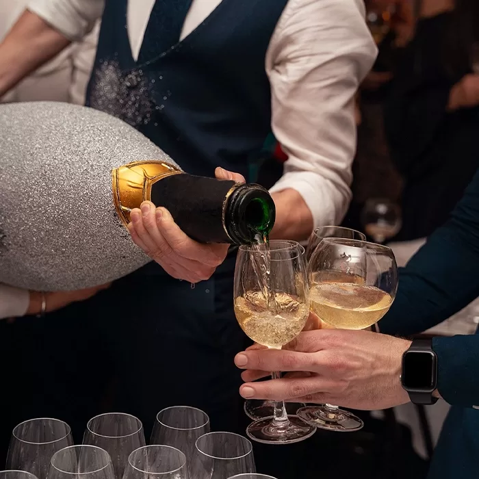 A person in formal attire pours champagne from a large, glittery bottle into two glasses held by another person at a celebratory event. Several empty glasses are visible on the table.