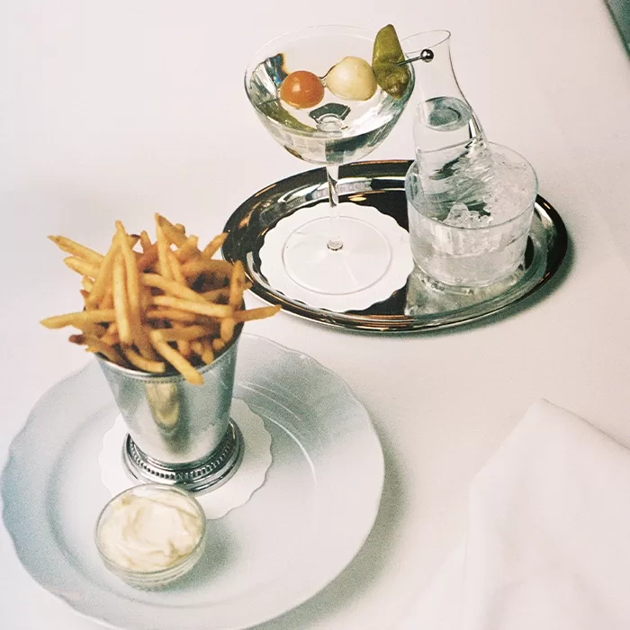 A metal cup of thin French fries with a side of dipping sauce sits on a plate. Nearby, a cocktail with garnishes and a carafe of water on a silver tray are arranged on a white tablecloth.