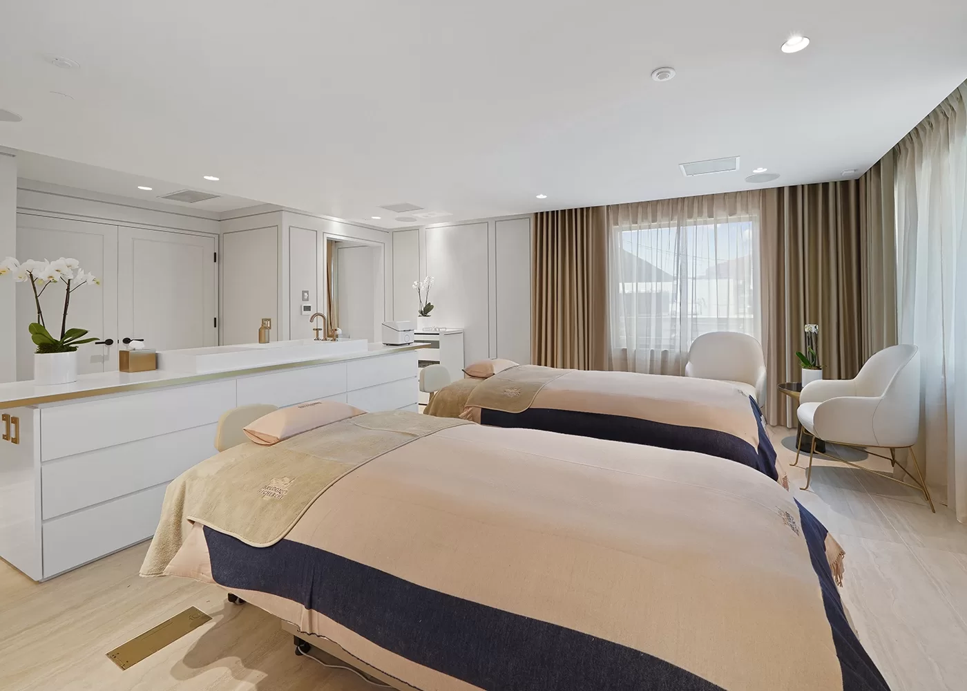 A modern, spacious spa room with two massage tables covered in beige and navy linens, white cabinetry with orchids, a large window with drapes, and two white armchairs in the corner. The room is bright and serene.