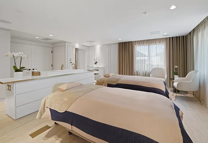 A bright, modern spa room with two beige massage tables, a white dresser with orchids, a chair by the window, light wood floors, and floor-to-ceiling curtains letting in natural light.