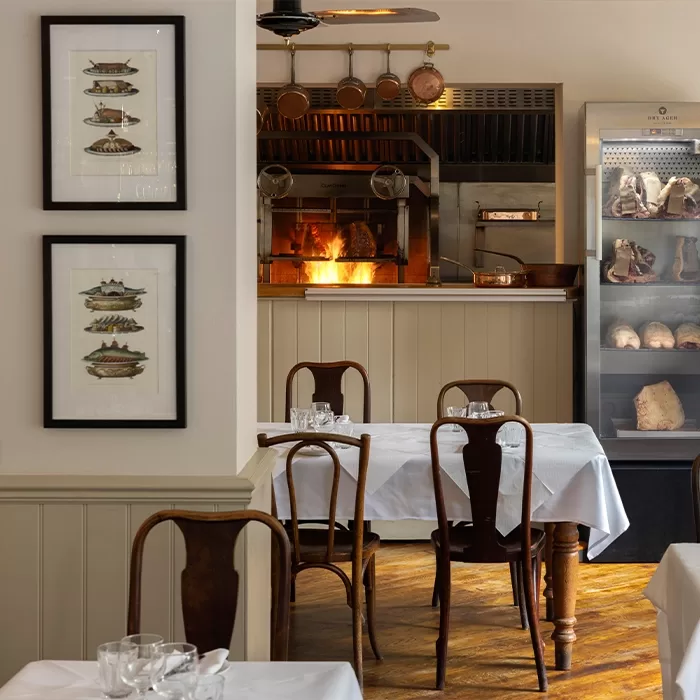 A cosy restaurant interior with white tablecloths, wooden chairs, framed food illustrations on the wall, an open kitchen with a wood-fired oven, and a glass meat refrigerator.