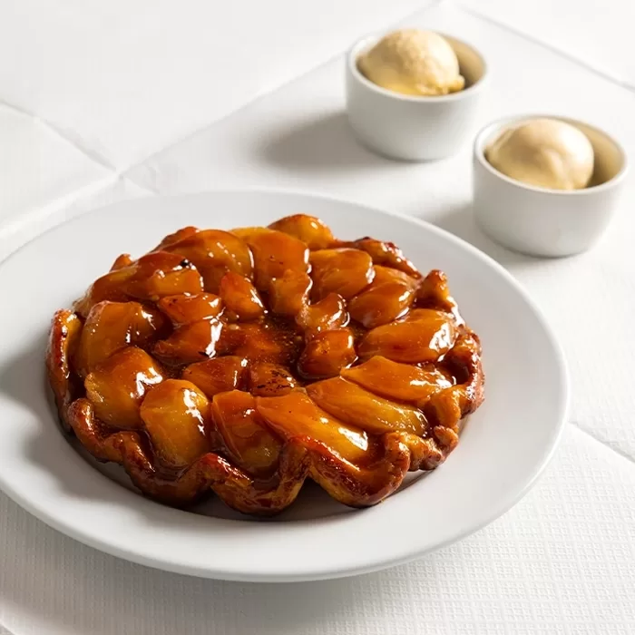A glazed apple tart sits on a white plate, with two small bowls of vanilla ice cream in the background, all placed on a white tablecloth.