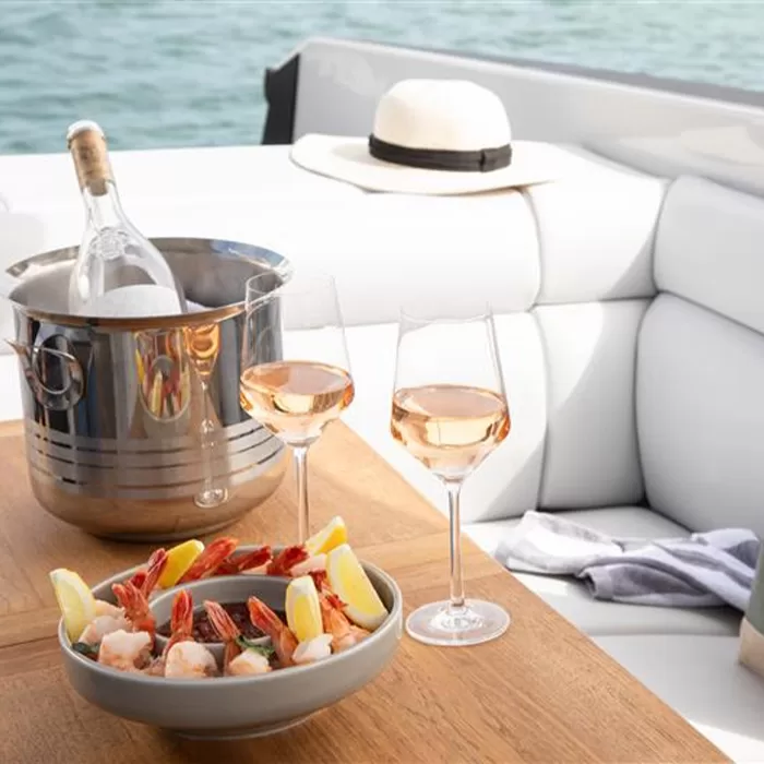 A bowl of shrimp with lemon wedges, two glasses of rosé wine, and a wine bottle in an ice bucket on a wooden table atop a white cushioned boat seat, with a white hat and water in the background.