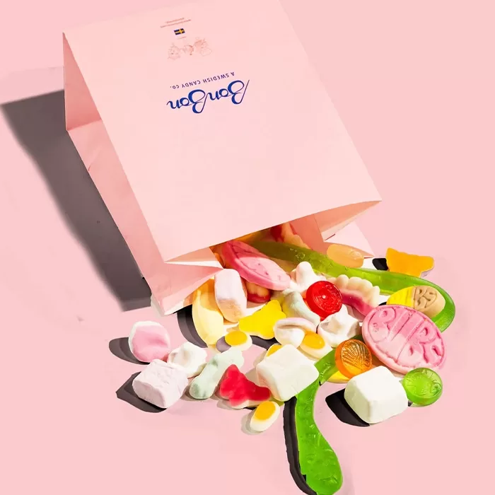 A pink paper bag labeled BonBon spills out assorted colorful candies onto a matching pink surface, including gummy eggs, fruit shapes, and marshmallows.