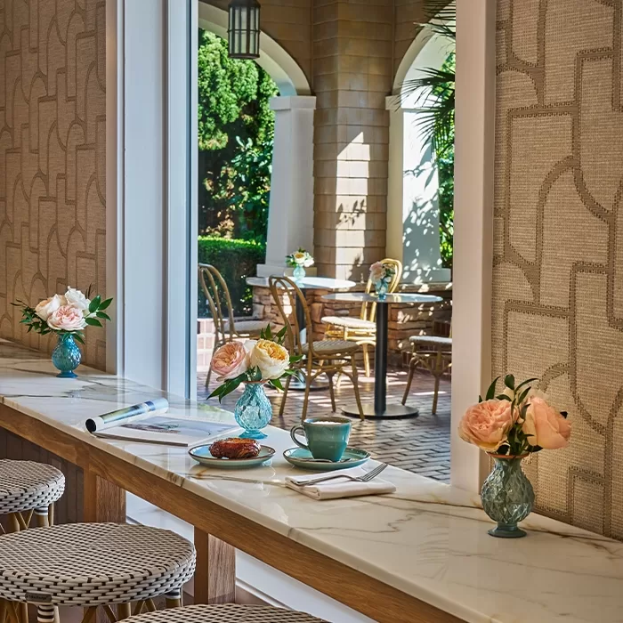 Cozy indoor café with marble countertop, blue cups and plates, pastries, and pink flower vases. Sunlit outdoor patio with chairs, tables, and green foliage visible through large window.