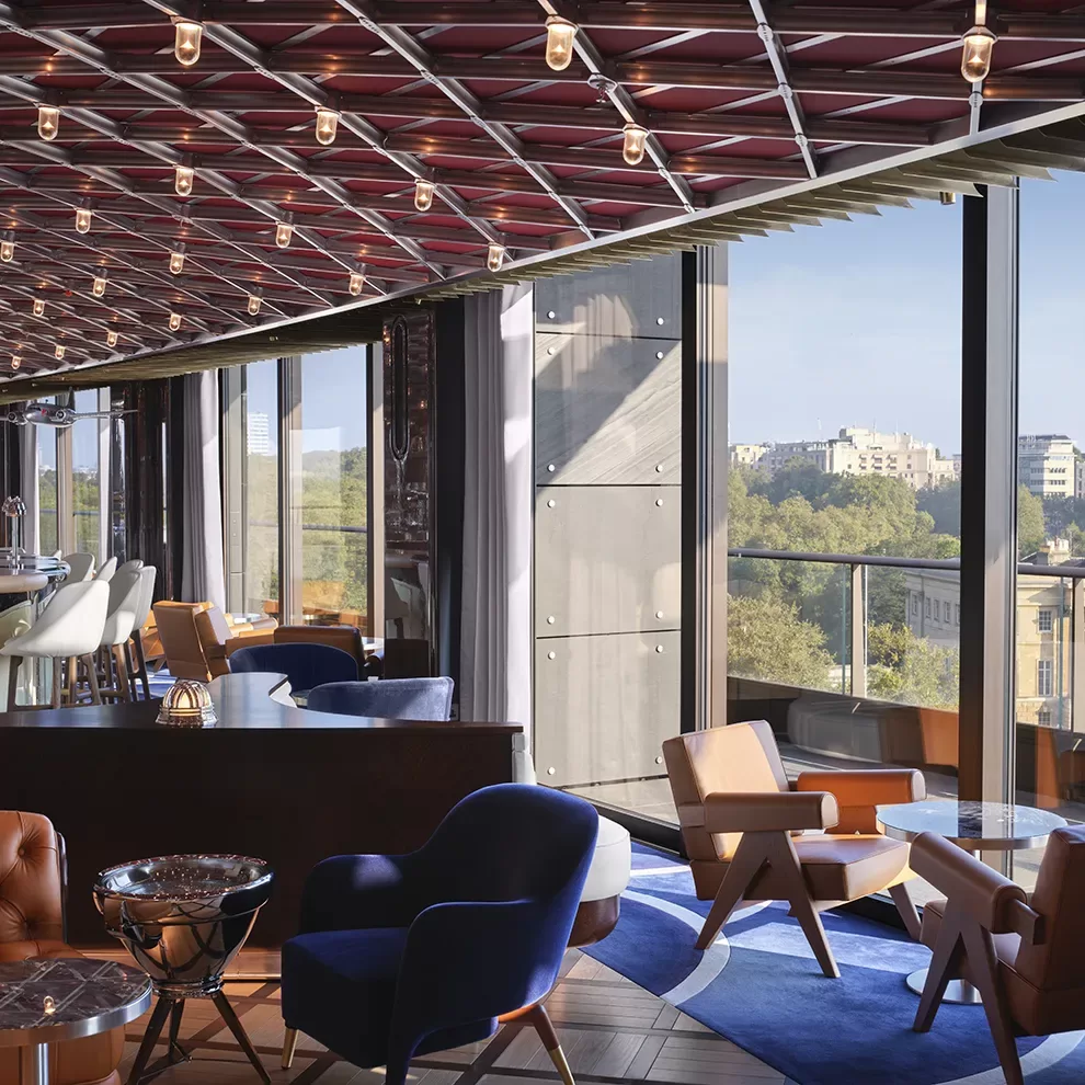 A modern lounge with floor-to-ceiling windows, offering a view of a cityscape. The room features various seating options, including blue and tan chairs, and a unique geometric ceiling with small lights. Natural light fills the space.