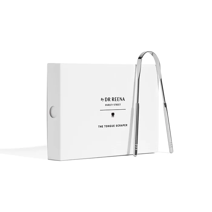 A sleek, curved metal tongue scraper leans against a white box labeled Dr Reena Harley Street. The minimalist packaging has a clean and modern design with a subtle tooth image.