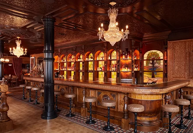 A luxurious bar with ornate wooden décor, arched shelves filled with bottles, cushioned bar stools, chandeliers, and stained glass accents, creating a warm, vintage atmosphere.
