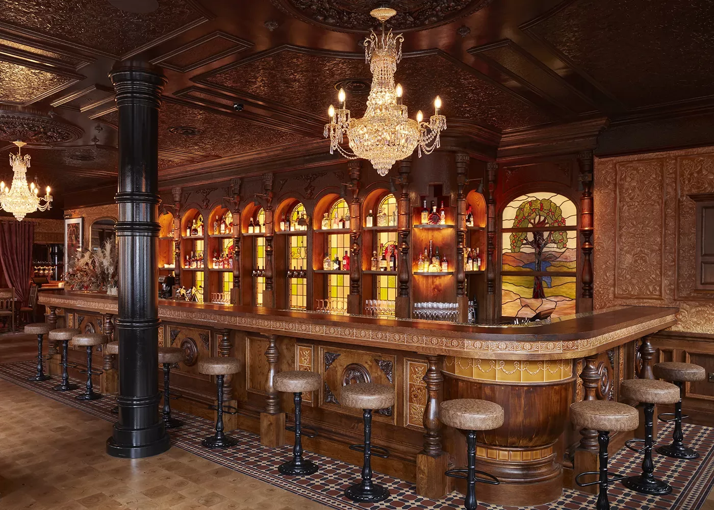 A luxurious, vintage-style bar with a wooden counter, padded barstools, a crystal chandelier, stained glass artwork, and shelves of bottles illuminated by warm lighting. Ornate patterns decorate the ceiling and walls.