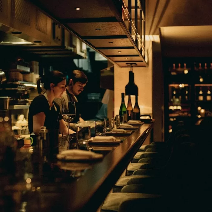 Dimly lit restaurant bar with two staff members preparing drinks behind the counter. Wine bottles, glasses, and plates are neatly arranged along the bar, creating a cozy, intimate atmosphere.