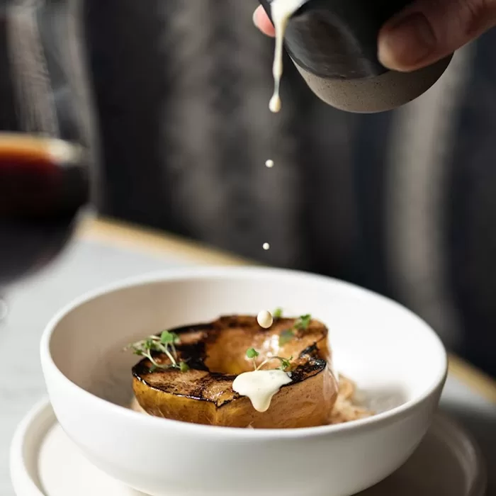 A hand pours creamy sauce over a roasted squash half garnished with microgreens, served in a white bowl on a matching plate.