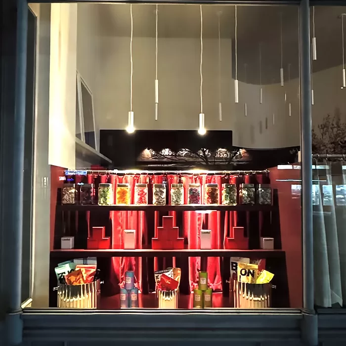A shop window display features jars of colorful candies on red shelves, with hanging pendant lights above. Packaged snacks are arranged on the lower shelves, creating a bright and inviting presentation.