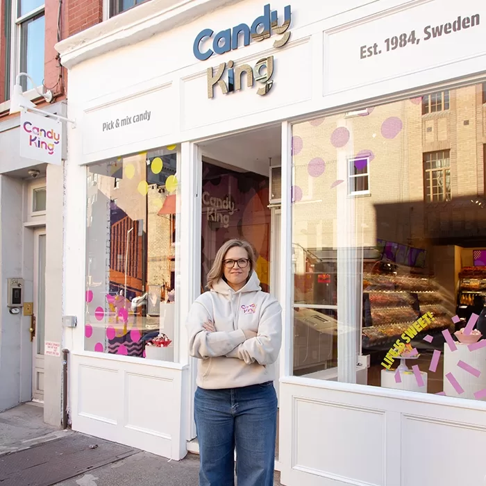 A person wearing glasses and a light hoodie stands with arms crossed in front of a white storefront labeled Candy King, featuring colorful candy displays in the window.