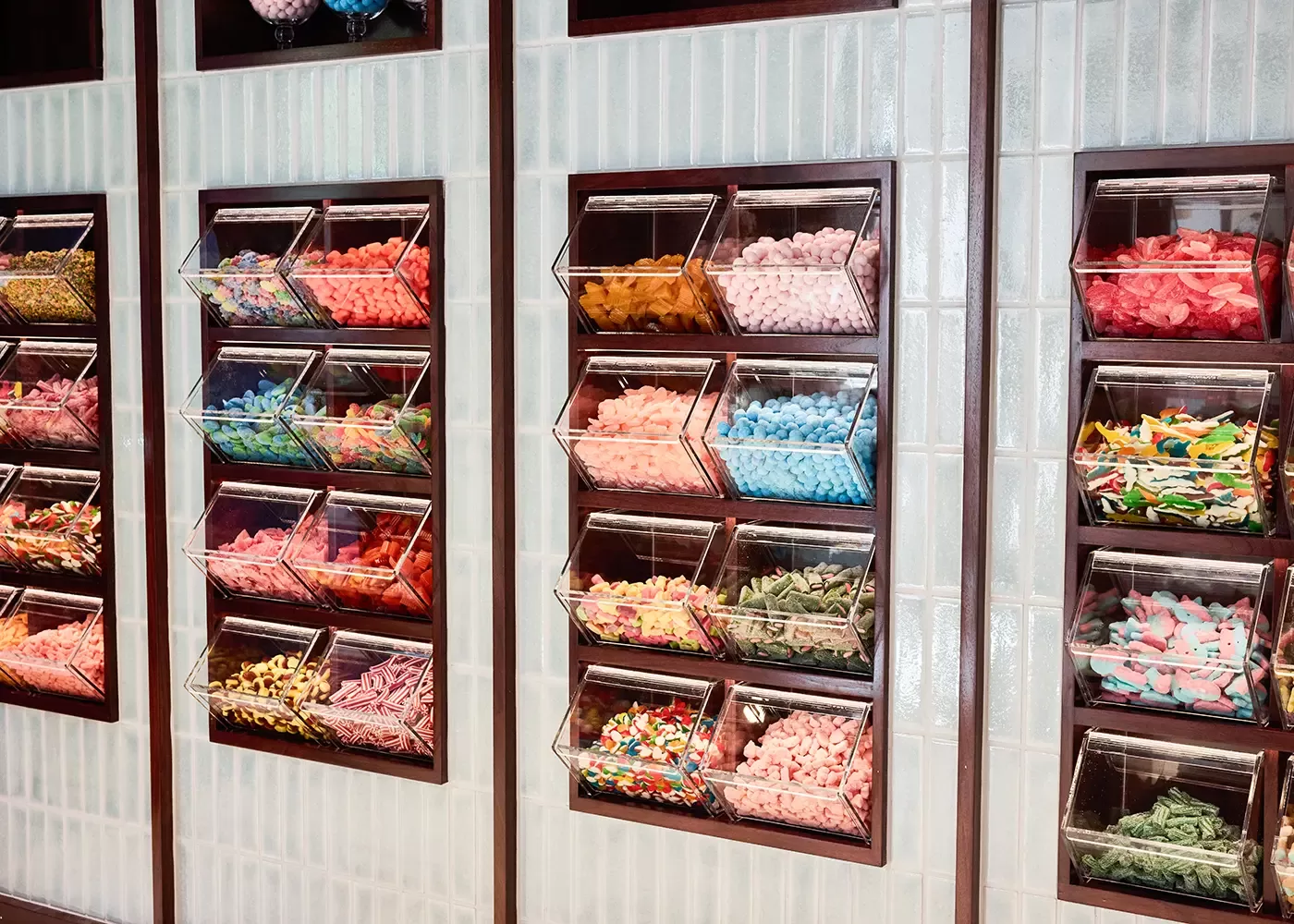 Clear plastic bins filled with various colorful candies, including gummies and licorice, are neatly arranged on a tiled wall in a self-serve candy shop.