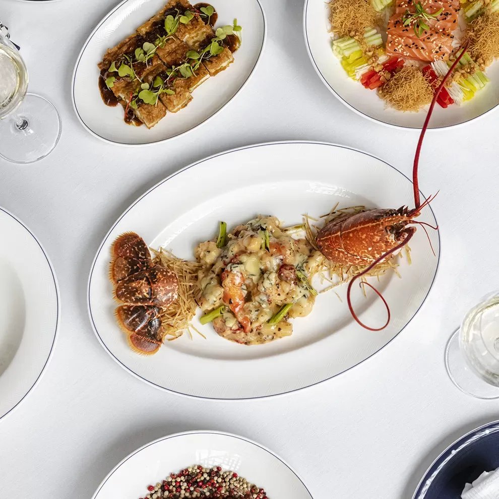 A white table setting with an oval plate featuring a cooked lobster, garnished with herbs. Surrounding dishes include stuffed rolls and a salmon dish. Glasses of white wine and a dish of peppercorns are nearby.