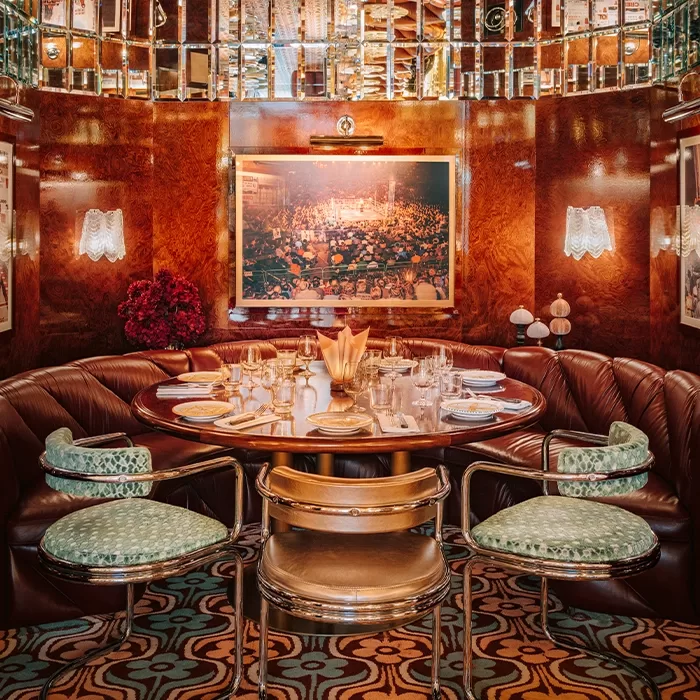 A cosy, upscale restaurant booth with brown leather seating, a round table set for dining, ornate chairs, a mirrored ceiling, rich wood-paneled walls, and elegant lighting, creating a warm and luxurious ambiance.