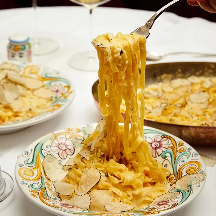 A fork lifts creamy tagliatelle pasta with shaved truffles from a decorative bowl, with more pasta and truffles on a plate and pan in the background, set on a white tablecloth.