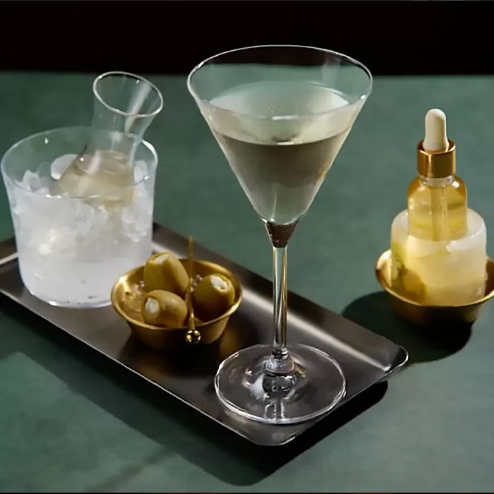 A martini glass with a clear cocktail sits on a tray next to a glass of ice with a small carafe, a gold dish of green olives on picks, and a gold bottle with a dropper, all on a dark green surface.