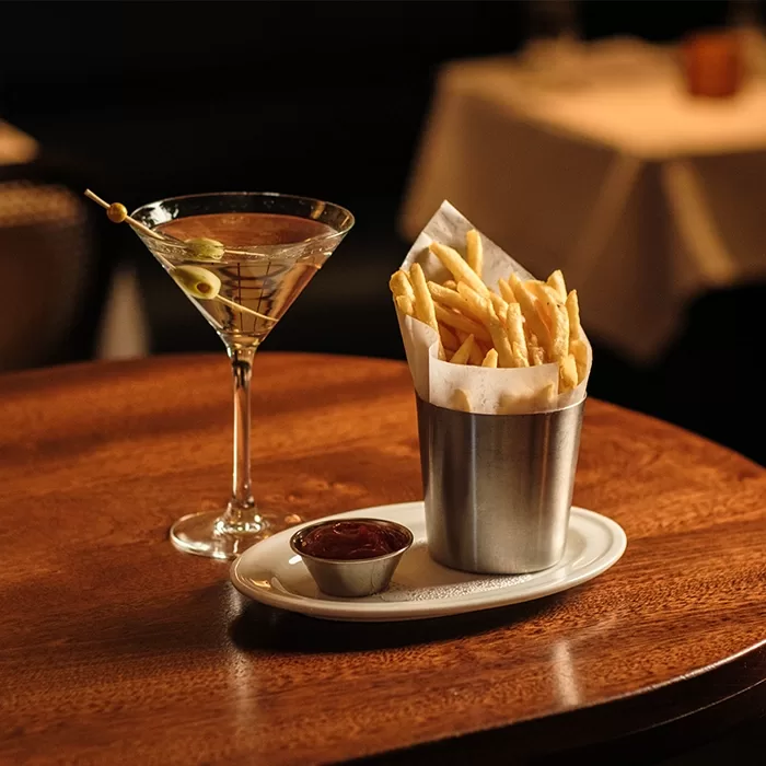 A glass of martini with olives on a cocktail pick sits beside a metal cup filled with French fries and a small dish of ketchup, all on a wooden table in a warmly lit restaurant.