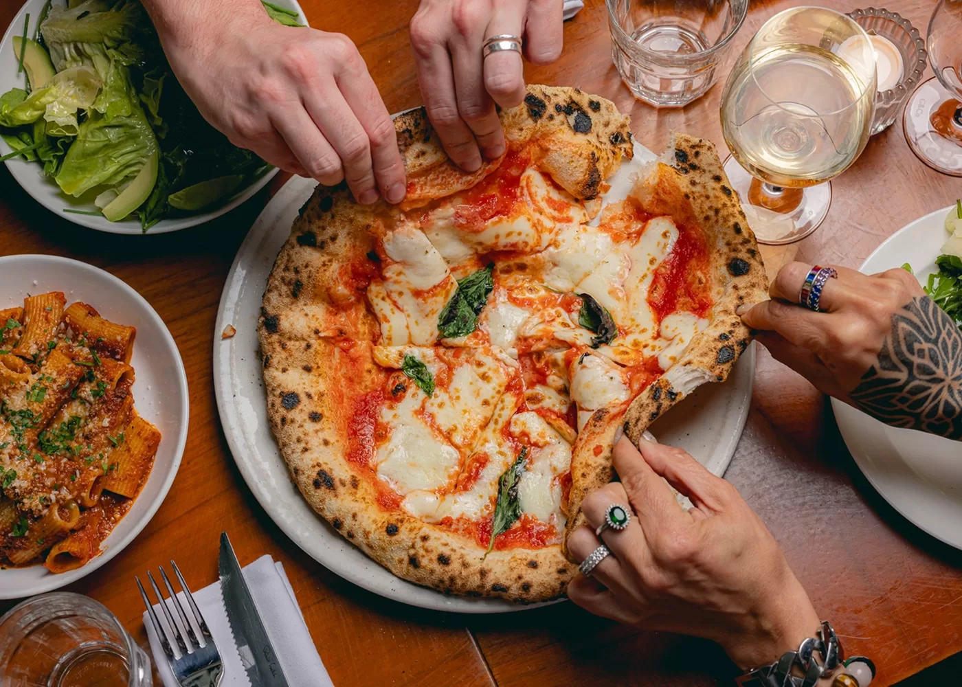 Three people reach for slices of a wood-fired pizza topped with mozzarella, basil, and tomato sauce on a wooden table set with pasta, salad, glasses, and cutlery.