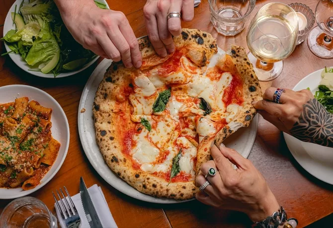Three hands reach for slices of a wood-fired margherita pizza on a table set with pasta, salad, wine, water glasses, and cutlery. The pizza has melted cheese, basil, and tomato sauce.