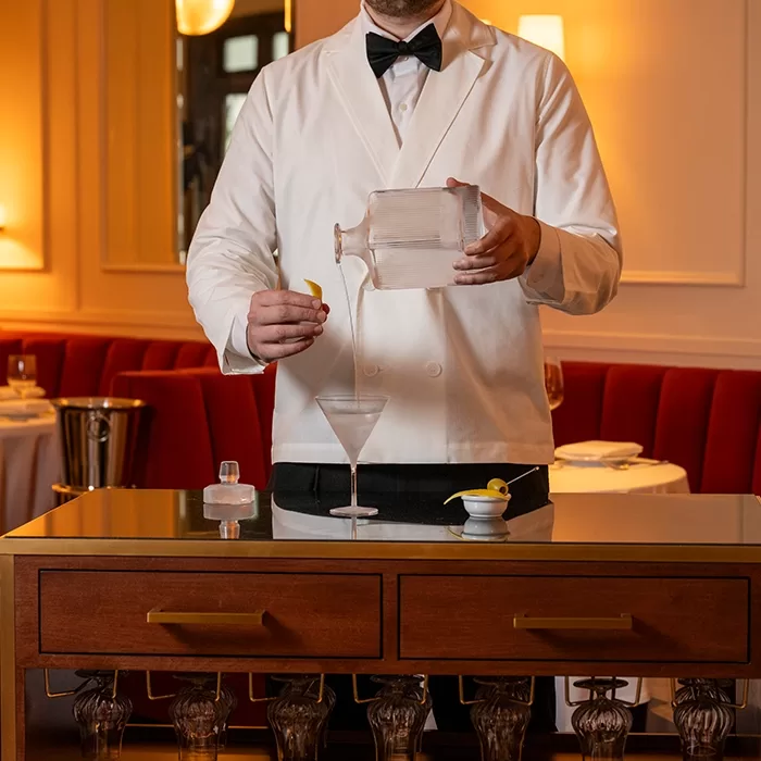 A waiter in a white jacket and bow tie pours a clear drink from a glass decanter into a martini glass, standing behind a wooden cart in an elegant restaurant with red velvet seating.