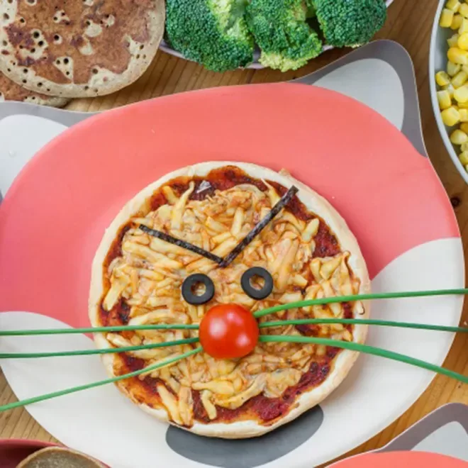 A childrens pizza arranged to look like a cat, with cheese, a cherry tomato nose, olive slices for eyes, and green chive whiskers. Its on a cat-themed plate, surrounded by broccoli, corn, and a tortilla.
