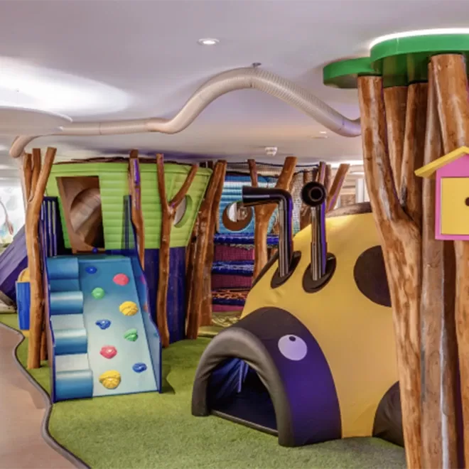 A colorful indoor playground features a slide with blue and green sections, climbing holds, and a tunnel shaped like a yellow and black bug. Wooden poles and a birdhouse decor add to the playful, nature-themed environment.