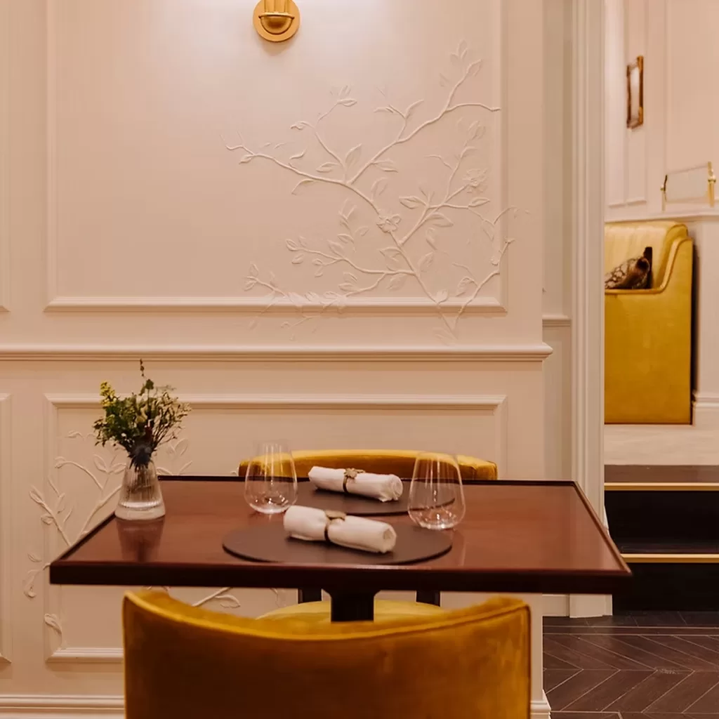A small elegant dining table set for two with rolled napkins and glasses, surrounded by mustard yellow velvet chairs, in a softly lit room with decorative wall molding and subtle floral designs.