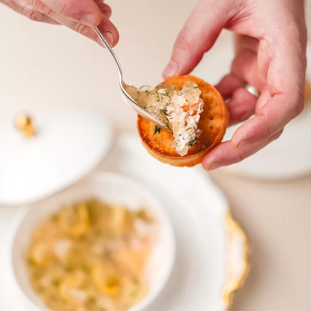 A person spreads a creamy, herb-filled topping onto a small round piece of toasted bread with a fork, above a white dish containing more of the topping.