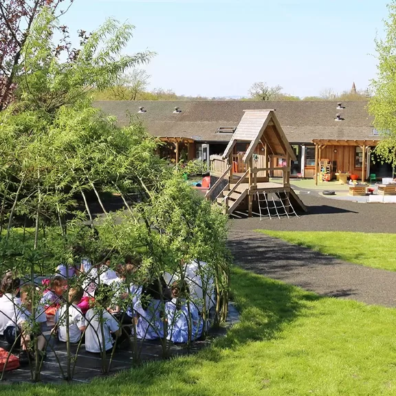 Children sit in a circular willow shelter on a grassy playground near a wooden climbing structure, with a school building and clear sky in the background.