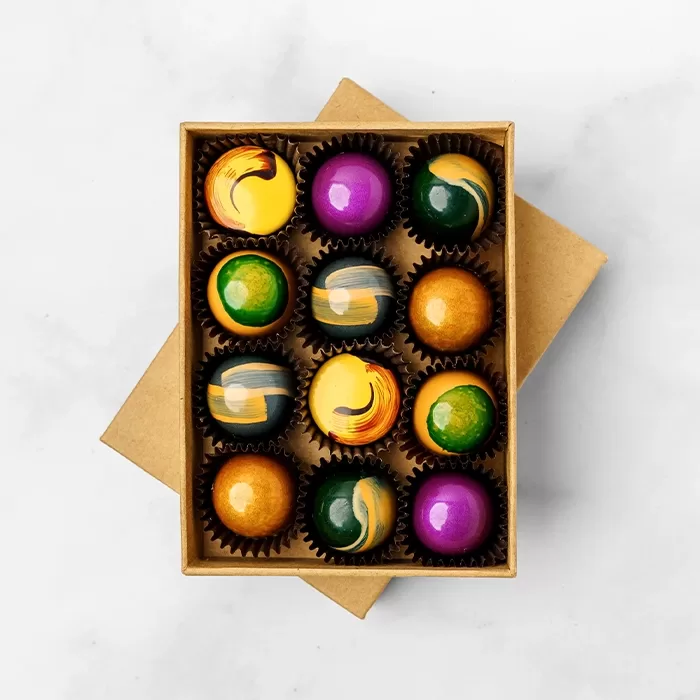 A box of twelve colorful, glossy chocolates with artistic designs in shades of yellow, green, purple, orange, and blue, arranged neatly in a kraft paper box on a white surface.