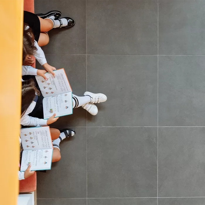 Two children in school uniforms sit on a bench reading books, with only their legs and books visible from a top-down view against a tiled floor.