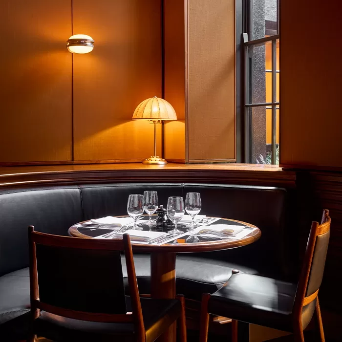 A cozy, warmly lit restaurant corner with a round table set for four, featuring wine glasses and napkins. Black leather seating and a table lamp create an inviting, intimate atmosphere near a window.