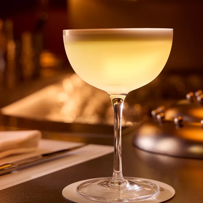 A coupe glass filled with a pale yellow cocktail sits on a white coaster on a dimly lit bar counter, with utensils and blurred background elements visible.